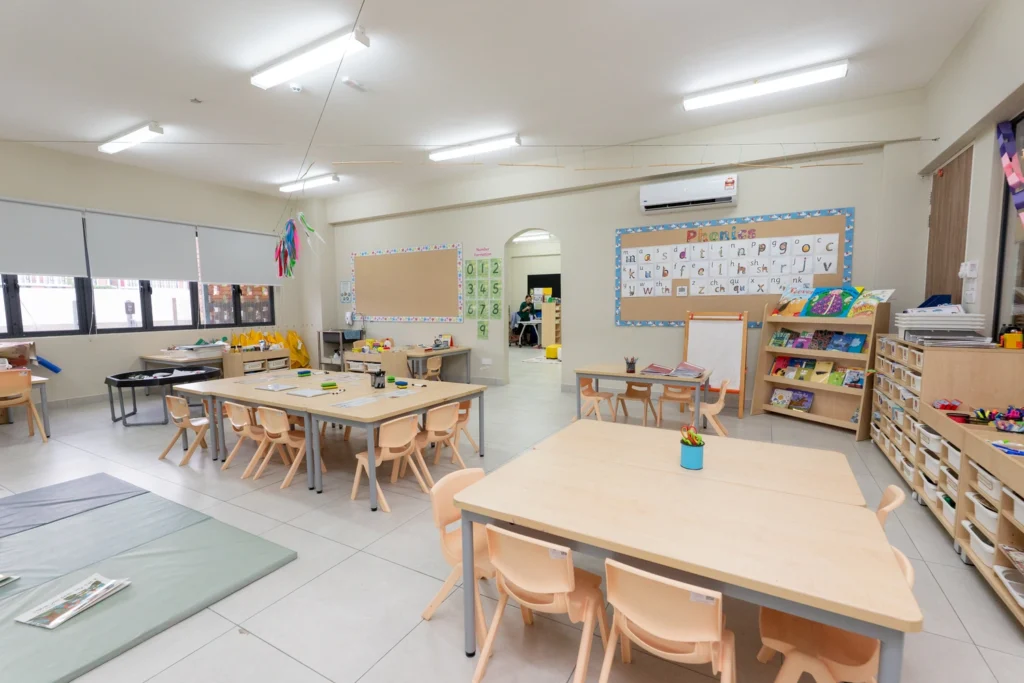 A bright early years classroom with wooden tables and chairs, educational materials on shelves, and colorful decorations.