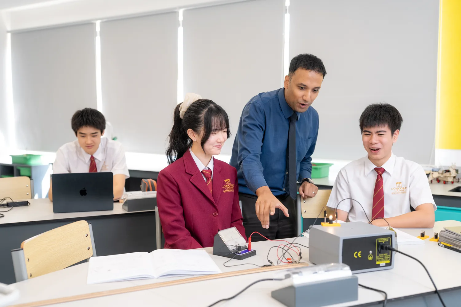 A physics teacher works with three Secondary students at a bench with electrical apparatus and a laptop.