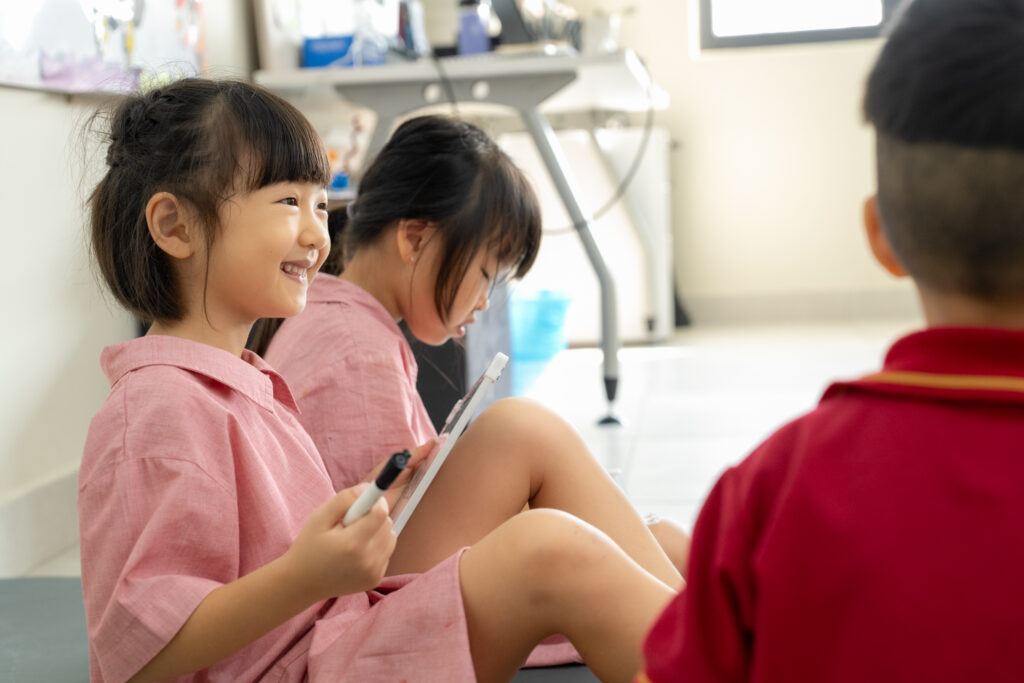Three students in pink uniforms are engaged with educational materials on the floor of a classroom.