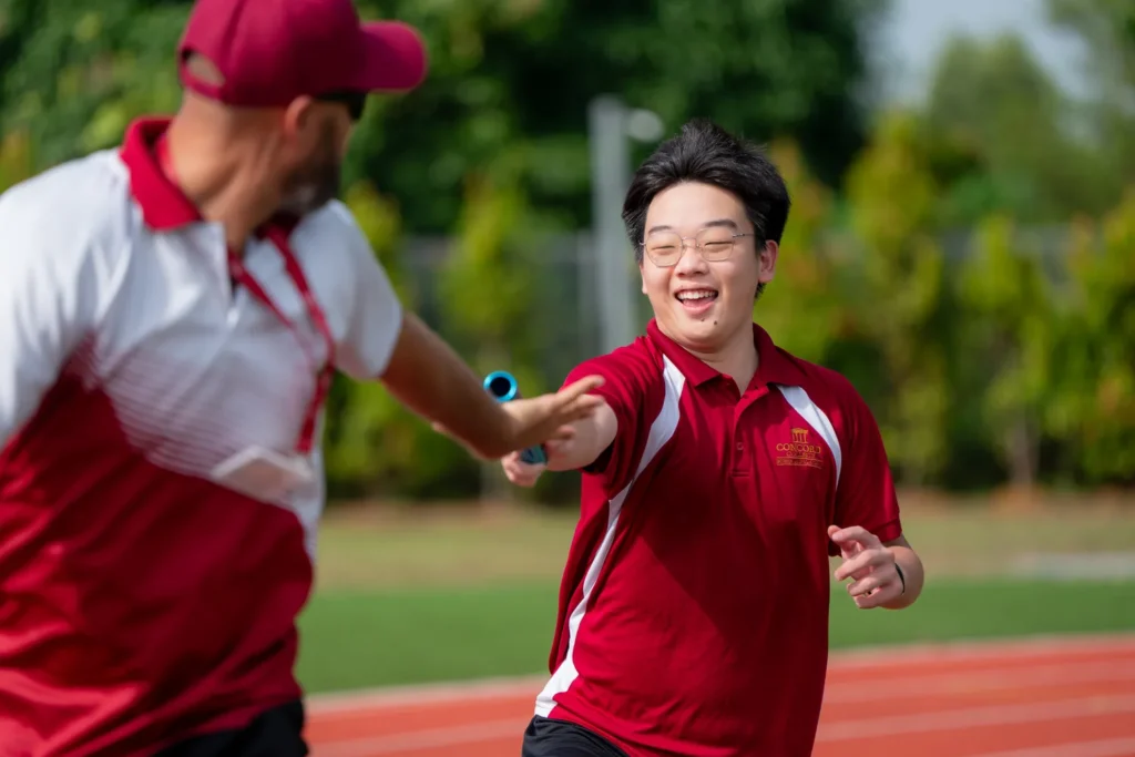 A coach passing a baton to a smiling student during a relay on the school track.