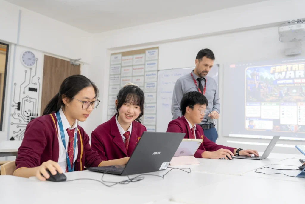 Students working at laptops in a computing lesson while their teacher looks on.