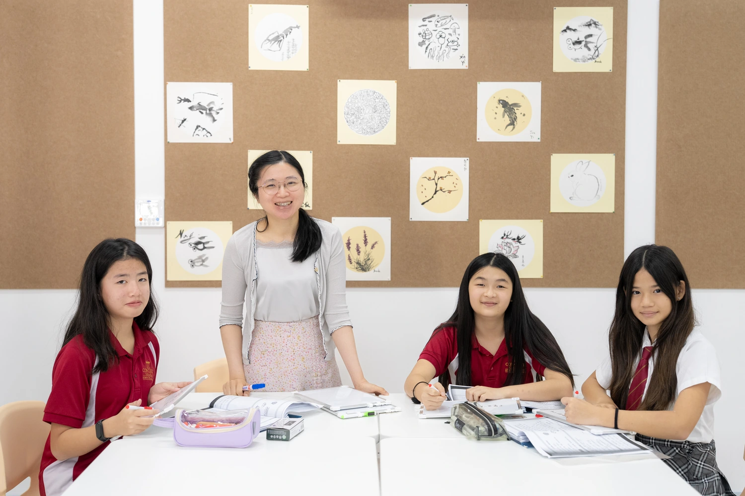 A teacher stands with three students at a table in the Mandarin classroom, Chinese brush paintings on the display board behind.