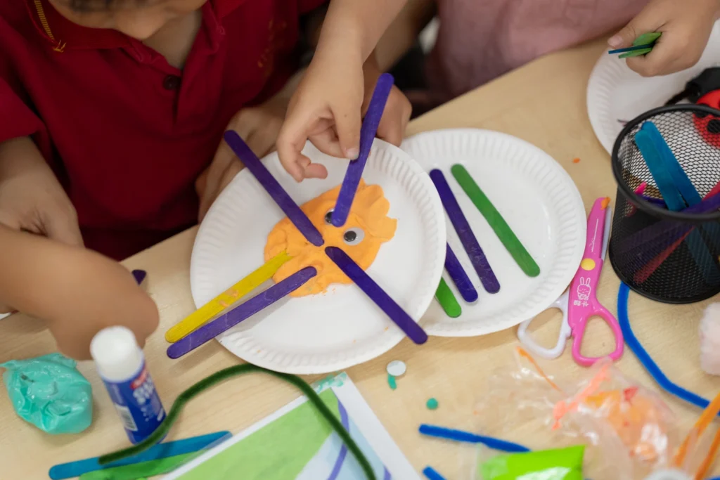 Children's hands glue coloured sticks onto a paper plate during a craft activity.