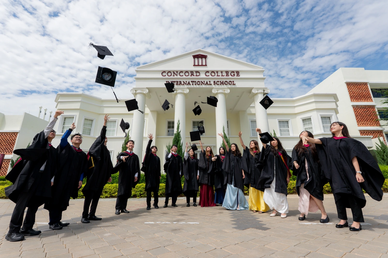 Year 13 graduates in gowns throwing their mortarboards into the air outside the Concord College International School building.