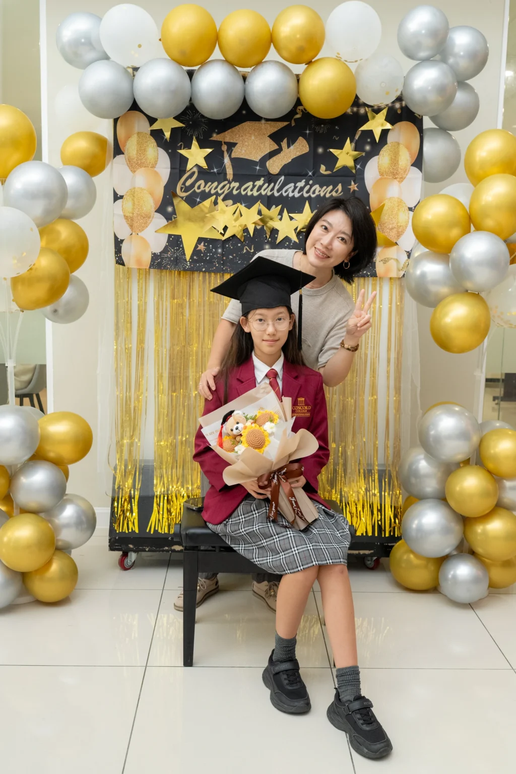 A student in a graduation gown holding a bouquet of flowers with a person standing behind them.